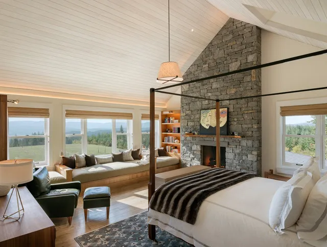 A four-poster bed shares a pale vaulted room with a stone fireplace and upholstered seating beside the windows.