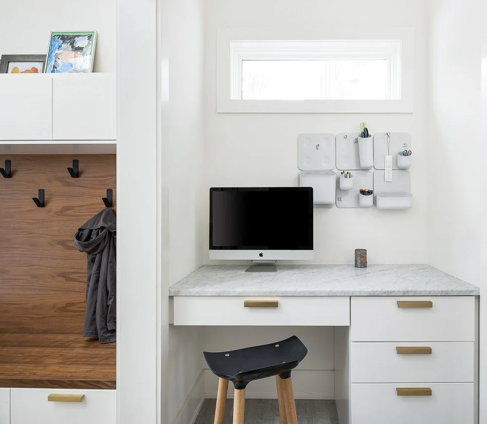 A white desk with drawers and a gray-veined countertop sits beneath wall organizers beside a wooden coat panel and a black stool