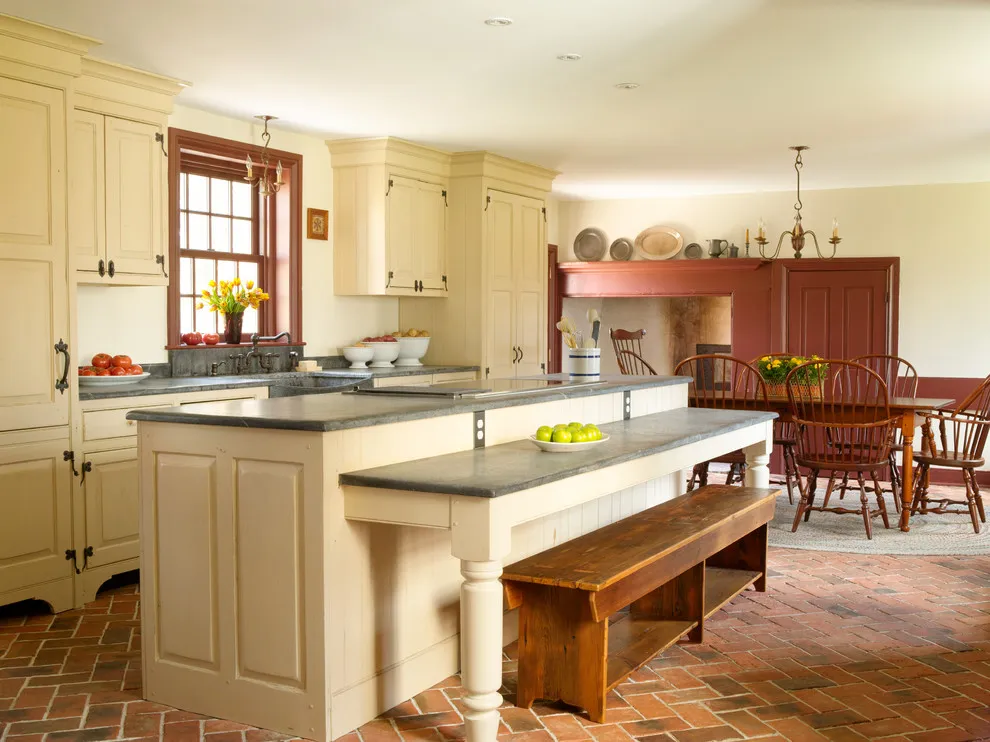 Cream cabinets and an island with a raised counter stand beside a wooden bench above a brick-patterned floor