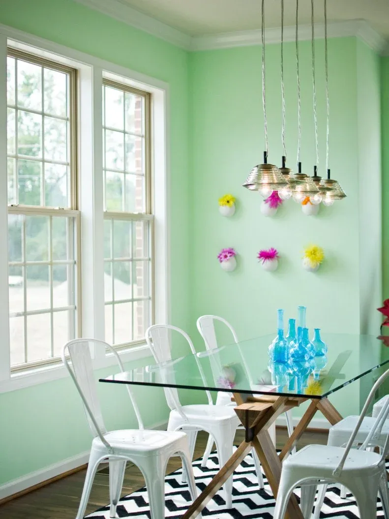 Clear glass table on a crossed wood-toned base with white chairs against mint-green walls and a zigzag rug