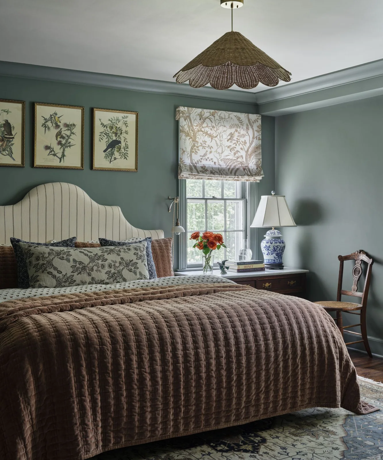 Muted teal walls surround a striped curved headboard, floral cushion, orange quilt, botanical pictures and a floral window blind
