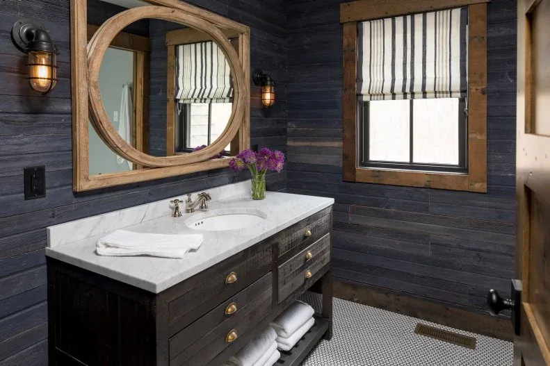 Dark blue boarded guest washroom with wood vanity, round wood mirror and striped window blind