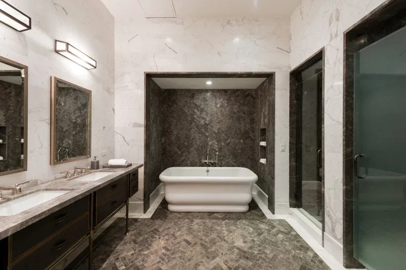 Pale freestanding bath in a brown-veined recess beside dark vanity storage and patterned flooring