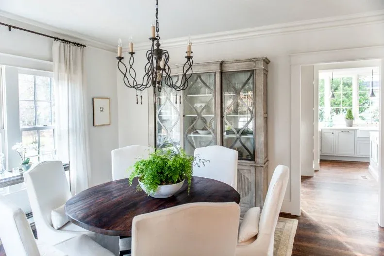 Dark open-arm chandelier with candle-shaped lamps above a round dark wooden table, pale upholstered chairs, glazed cabinets, and bright windows.