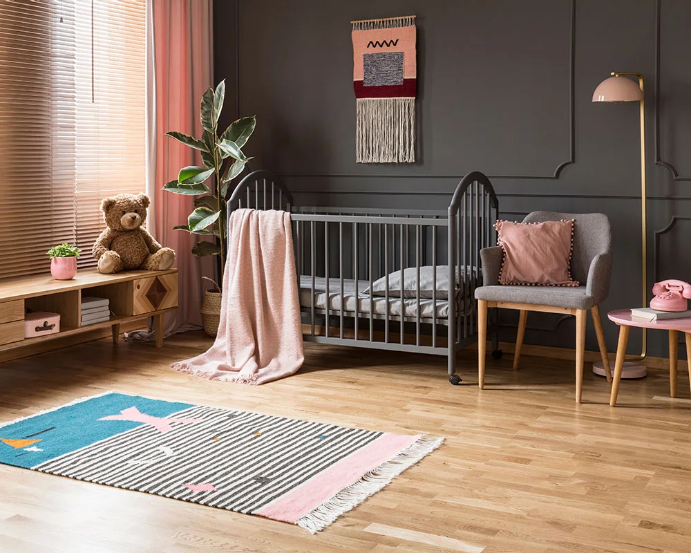 A dark nursery crib and pale upholstered chair sit beside a charcoal wall, pink curtains, a low wood cabinet and a small colorful rug