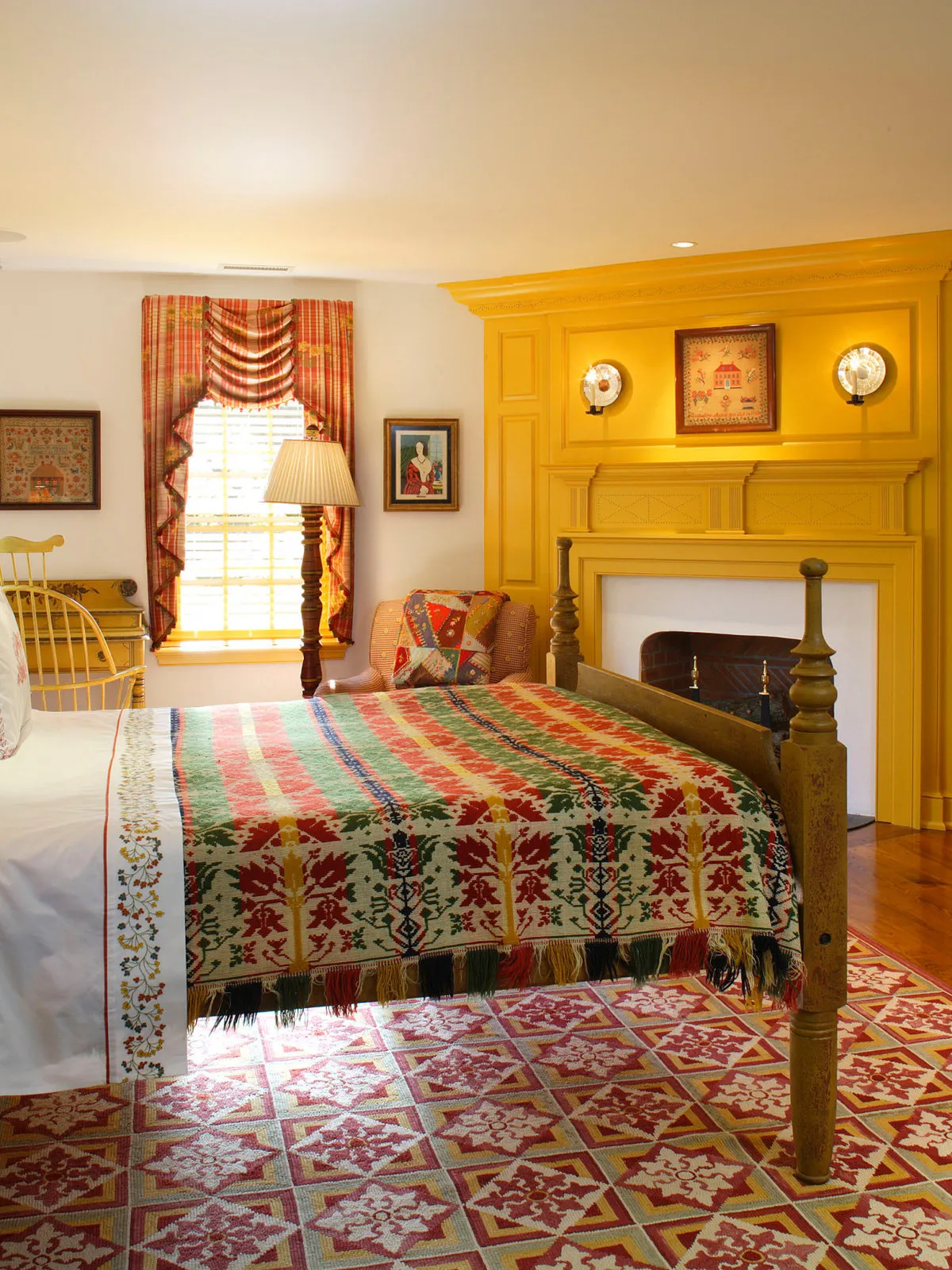 A colorful fringed coverlet dresses a bed beside a yellow mantel and white walls.