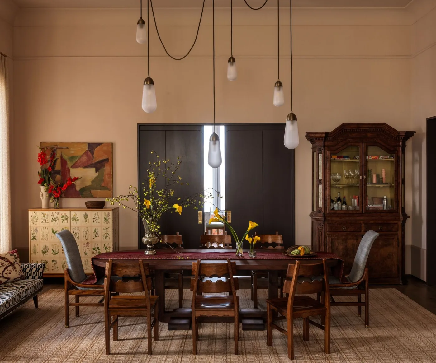Warm dining room with pale peach walls, wooden rectangular table, mixed chairs, a dark glass-front cupboard and several hanging small lights.