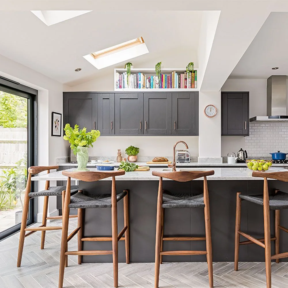 Gray kitchen cabinets and an island sit below white sloped ceilings and skylights beside wood-framed stools and pale subway tile