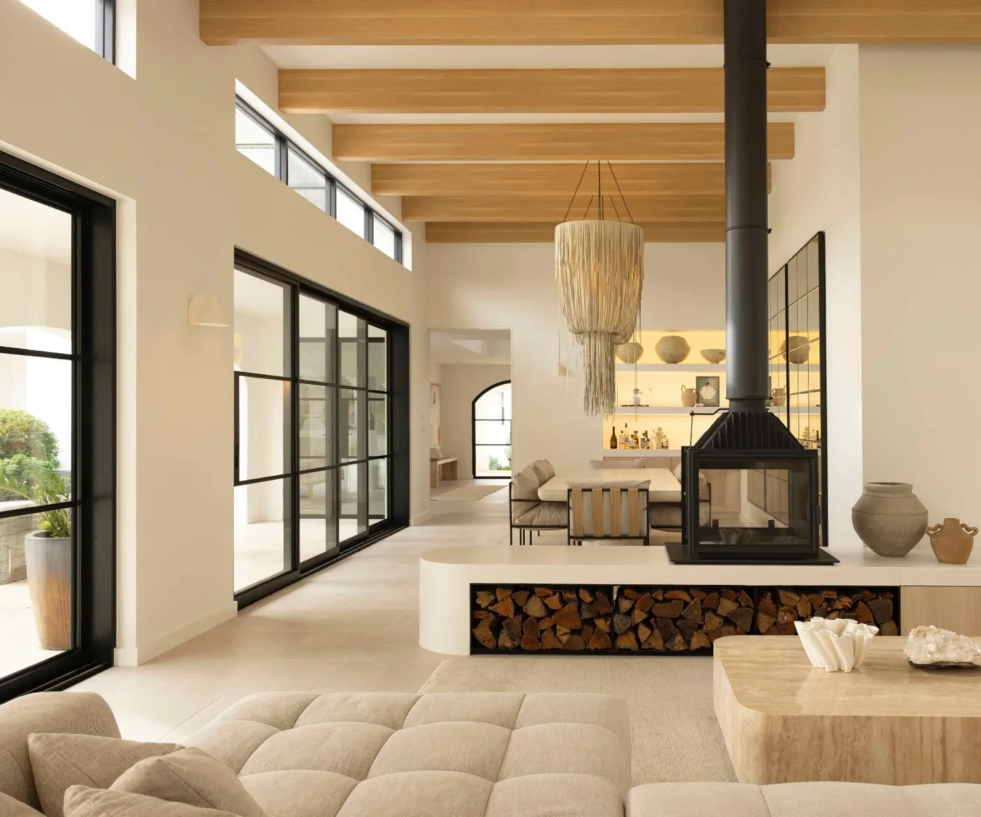 An open pale living room combines warm ceiling beams, large black-framed glazing, textured seating and a freestanding black fireplace above a log-storage ledge