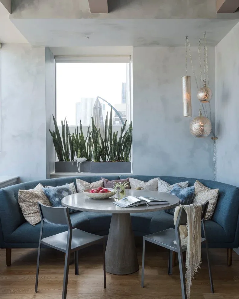 Curved blue upholstered banquette around a round gray table, with gray chairs, mixed pillows, plants by a window, and three hanging lights against pale walls.