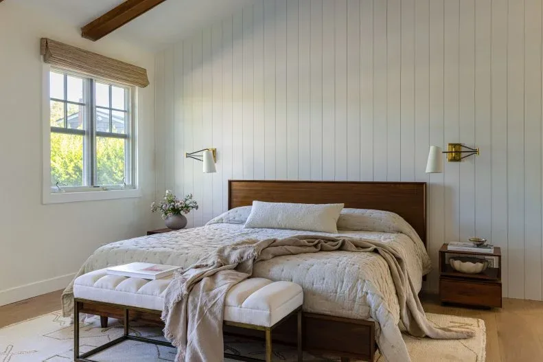 White vaulted bedroom with vertical wall boards, low wood headboard, bedside sconces and pale bench