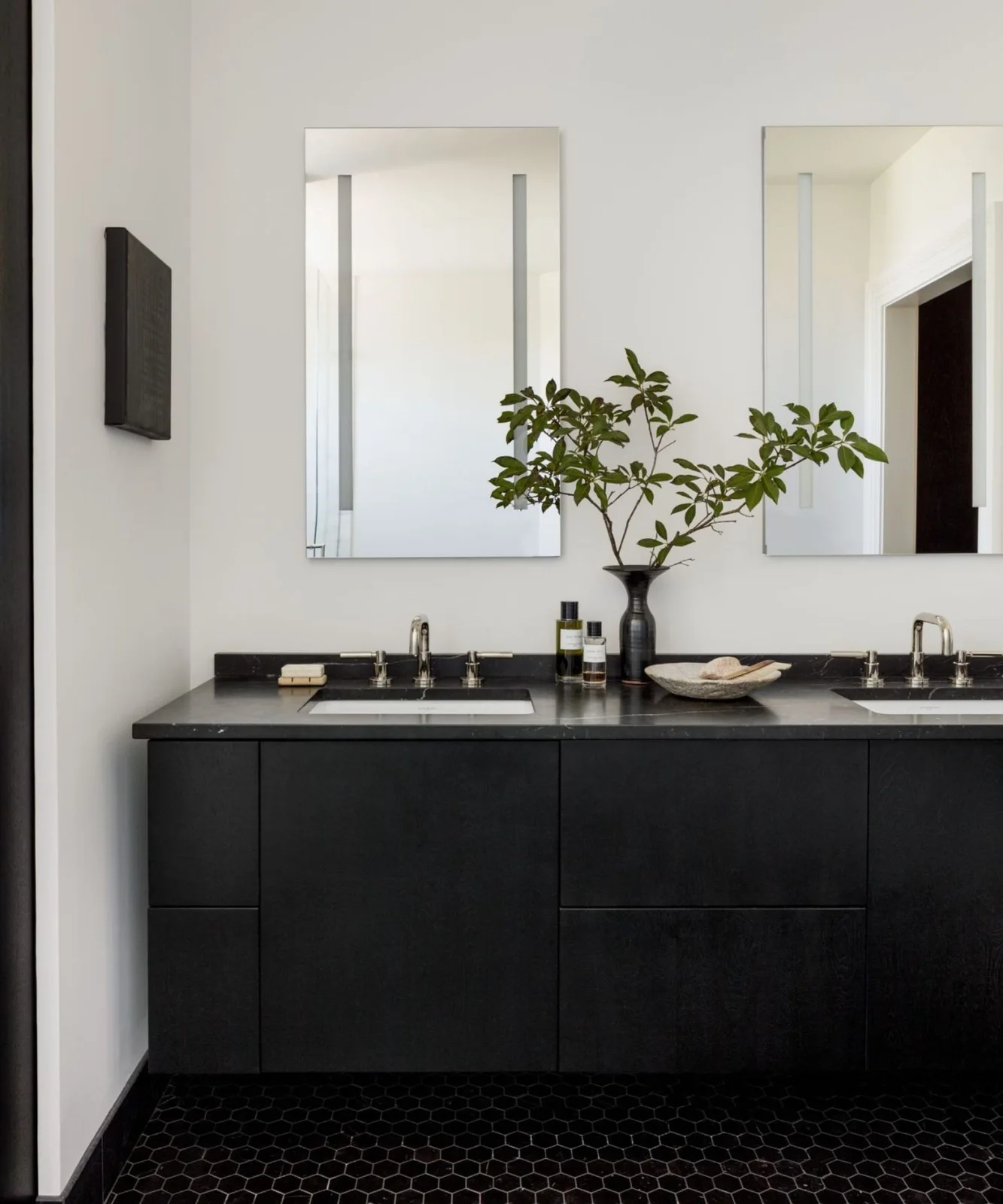 A black double vanity with a dark counter stands beneath two narrow rectangular mirrors beside pale walls and a small black hexagonal tiled floor