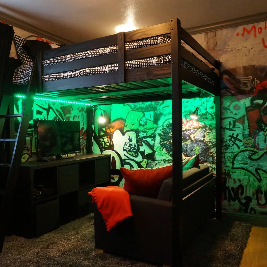 A dark loft bed stands above a desk and monitor beside a graffiti-covered wall and chair, with green accent lighting below the frame